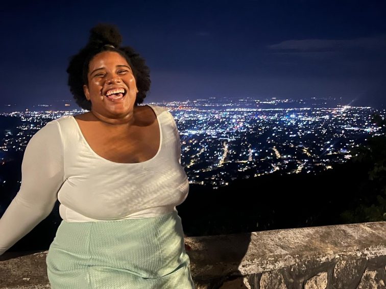 Picture of Samantha in a white long sleeve top tucked into a green shirt with the skyline in the background at night with lots of twinkling lights