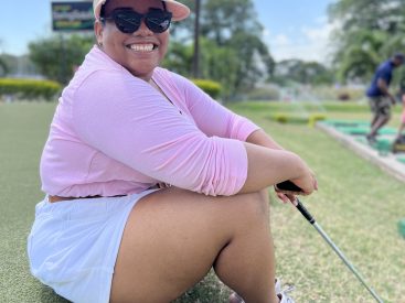 Samantha sitting on a golf course smiling in a pink hate, shades, pink long sleeve shirt tucked into white shorts and pink sneakers.