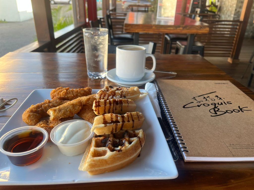 Picture of chocolate chip waffles with chicken tenders, a cup of ice water and coffee with a sketch book to the side on an outdoor table at a cafe
